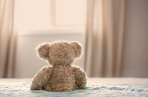 A teddy bear sitting on a bed looking out a window in soft light
