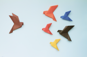 One paper crane turned towards a small group of multicolor paper cranes on a light blue background