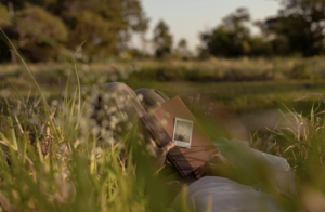 The legs of a woman resting in a field with a book and a polaroid of flowers resting on her lap